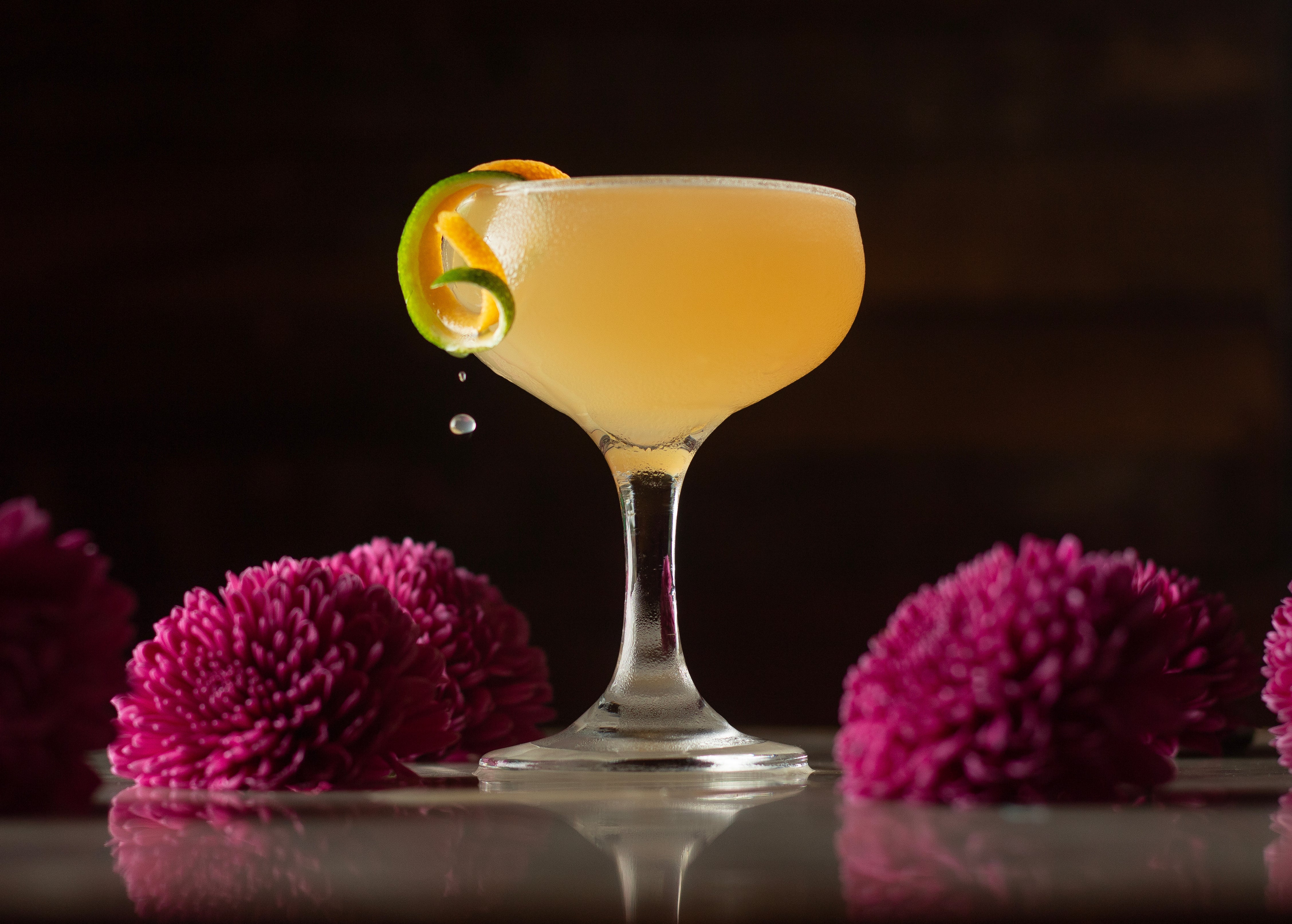Cocktail in a glass with a lime wheel garnish, surrounded by purple flowers on a dark background