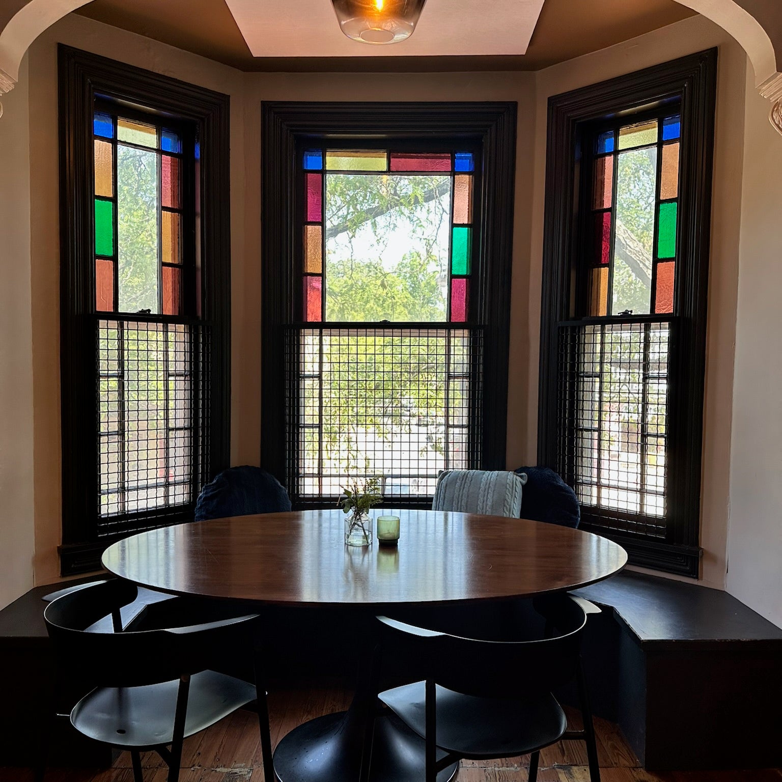 Dining room with round table and chairs in front of stained glass windows.